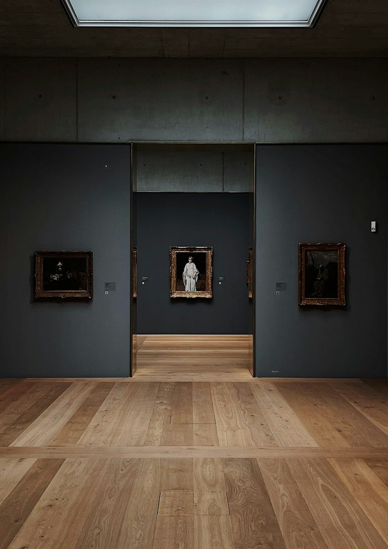 Ein neutrales Kunstmuseum mit dunklen Wänden und Holzboden, Blick auf Gemälde und Skulpturen.